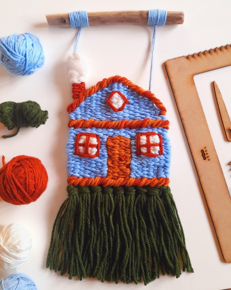 May include: A handmade woven wall hanging in the shape of a house. The house is blue with orange trim, a white chimney, and green fringe. Blue yarn is used for the hanging cord. Additional yarn balls in blue, orange, green, and white are scattered around the house.