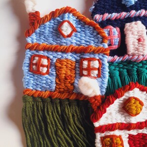 May include: Handmade textile art featuring colorful, whimsical houses. The houses are crafted with blue, red, white, and orange yarn, with green fringe at the base. Each house has unique details, including windows, doors, and chimneys.