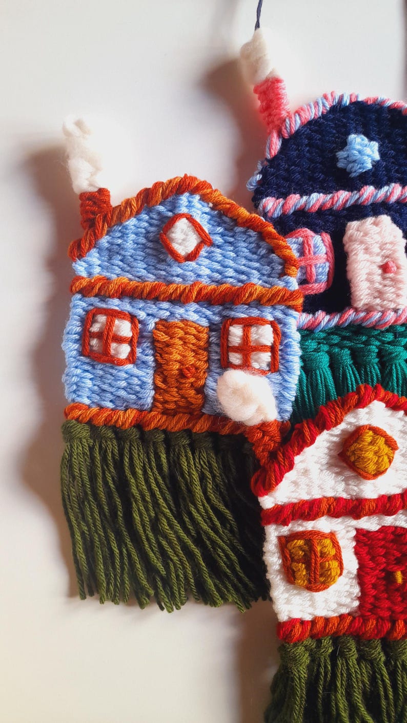 May include: Handmade textile art featuring colorful, whimsical houses. The houses are crafted with blue, red, white, and orange yarn, with green fringe at the base. Each house has unique details, including windows, doors, and chimneys.