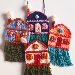 May include: Handmade textile wall hangings featuring colorful woven houses with fringed bottoms. The houses are in shades of blue, orange, red, and white, with details like windows, doors, and chimneys. They are suspended from a wooden branch.