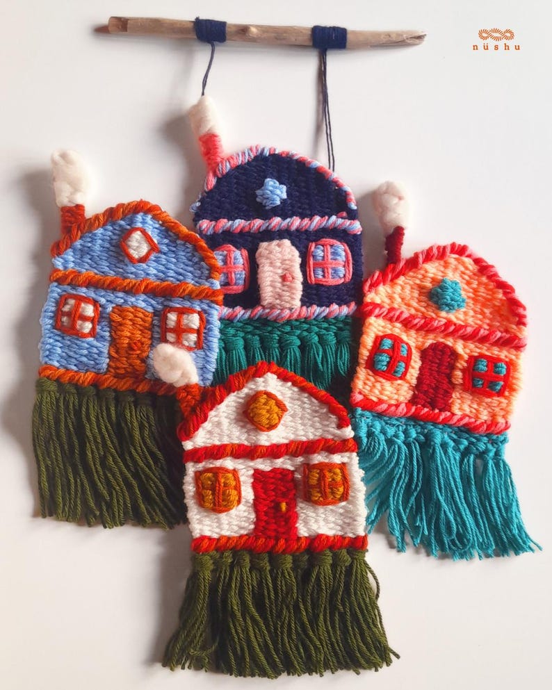 May include: Handmade textile wall hangings featuring colorful woven houses with fringed bottoms. The houses are in shades of blue, orange, red, and white, with details like windows, doors, and chimneys. They are suspended from a wooden branch.