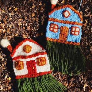 May include: Two handmade, woven house ornaments. One is blue with orange trim, a brown door, and green fringe. The other is white with red trim and door, and green fringe. Both have chimneys and windows.
