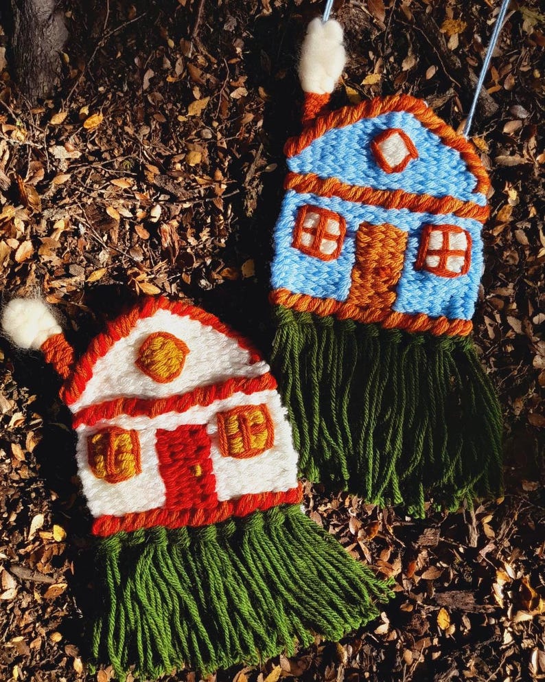May include: Two handmade, woven house ornaments. One is blue with orange trim, a brown door, and green fringe. The other is white with red trim and door, and green fringe. Both have chimneys and windows.