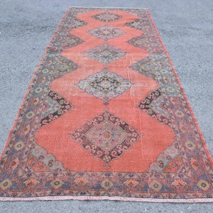 May include: A rectangular rug with a distressed appearance, featuring a coral-coloured central field and detailed geometric patterns in blue, brown, and black. The rug has a diamond design and decorative borders.