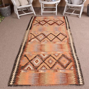 Könnte beinhalten: Ein rechteckiger, handgewebter Teppich mit geometrischem Muster. Der Teppich hat ein zentrales Design aus Rauten und Zickzack in Orange-, Braun- und Beigetönen. Die Ränder sind mit einem schwarz-weißen Zickzackmuster eingefasst.