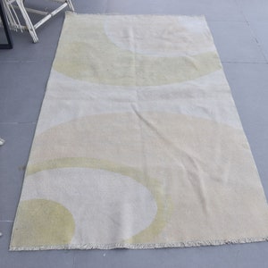May include: A rectangular rug with a cream-colored background and large, overlapping abstract shapes in light green and beige. The rug has a fringed edge and is placed on a gray surface.