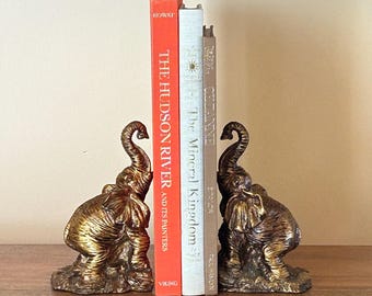 May include: A pair of gold elephant bookends holding up three books. The books are titled "The Hudson River and Its Painters" by Howat, "The Mineral Kingdom" by Anne Taylor, and "Celanne" by Paul Salmon.