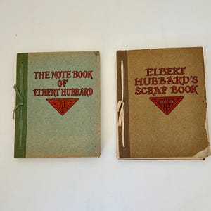 May include: Two vintage books: "The Note Book of Elbert Hubbard" and "Elbert Hubbard's Scrap Book." The books have decorative red and brown covers with text. The first book is light green with a green tie, and the second is brown with a tan tie.