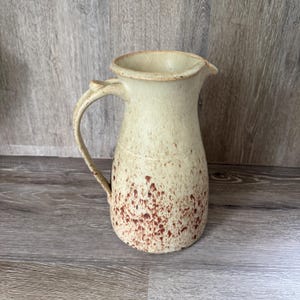 May include: A ceramic pitcher with a wide, flared rim and a curved handle. The body features a cream-colored base with a dense, speckled pattern of reddish-brown spots that become more concentrated toward the bottom of the vessel.