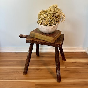 May include: A rustic, three-legged wooden stool with a small handle. A vintage book and a white bowl filled with dried flowers sit on top. The stool is stained a dark brown and shows signs of wear.