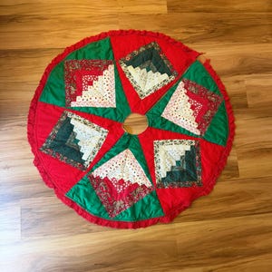 May include: A circular Christmas tree skirt featuring a patchwork design with alternating red and green fabric panels. Each panel contains a square log cabin quilt block made of various patterned fabrics in shades of red, green, and cream.