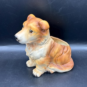 May include: A ceramic dog planter in shades of brown, tan, and white. The dog is sitting with its head turned to the side, and has a planter opening on its back. The dog has a glossy finish and is set against a dark background.