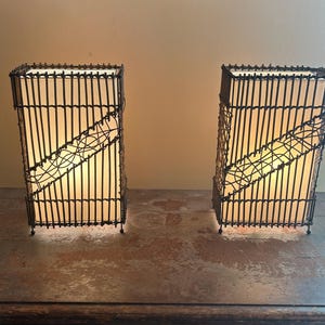 May include: Two rectangular lamps with a woven, dark brown frame and a cream-colored interior. The lamps have a diagonal, decorative wire design. They are placed on a wooden surface against a neutral background.