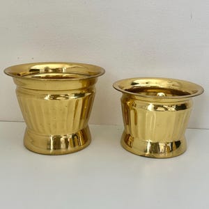 May include: Two gold metal planters with a ribbed design. The larger planter is taller and wider than the smaller planter.