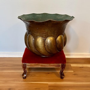 May include: A large, antique bronze-colored planter with a textured surface and a scalloped rim. The planter sits on a small, red velvet-covered stool with ornate legs. The interior of the planter is a dark green color.