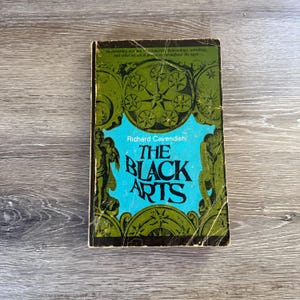 May include: A paperback book titled The Black Arts by Richard Cavendish with a bright blue center panel and olive green borders. The cover features black illustrations of circular star patterns and figures, with text at the top and center.