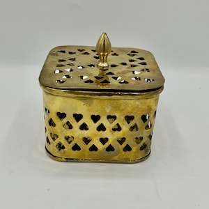 May include: A vintage brass box with a lid. The box is square with heart-shaped cutouts on the sides and a decorative finial on top. The lid also has cutouts. The brass has a warm, golden color.
