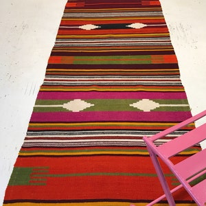 May include: A long, colorful rug with a striped pattern and geometric designs in shades of red, orange, green, pink, and white. The rug has fringe on the ends.