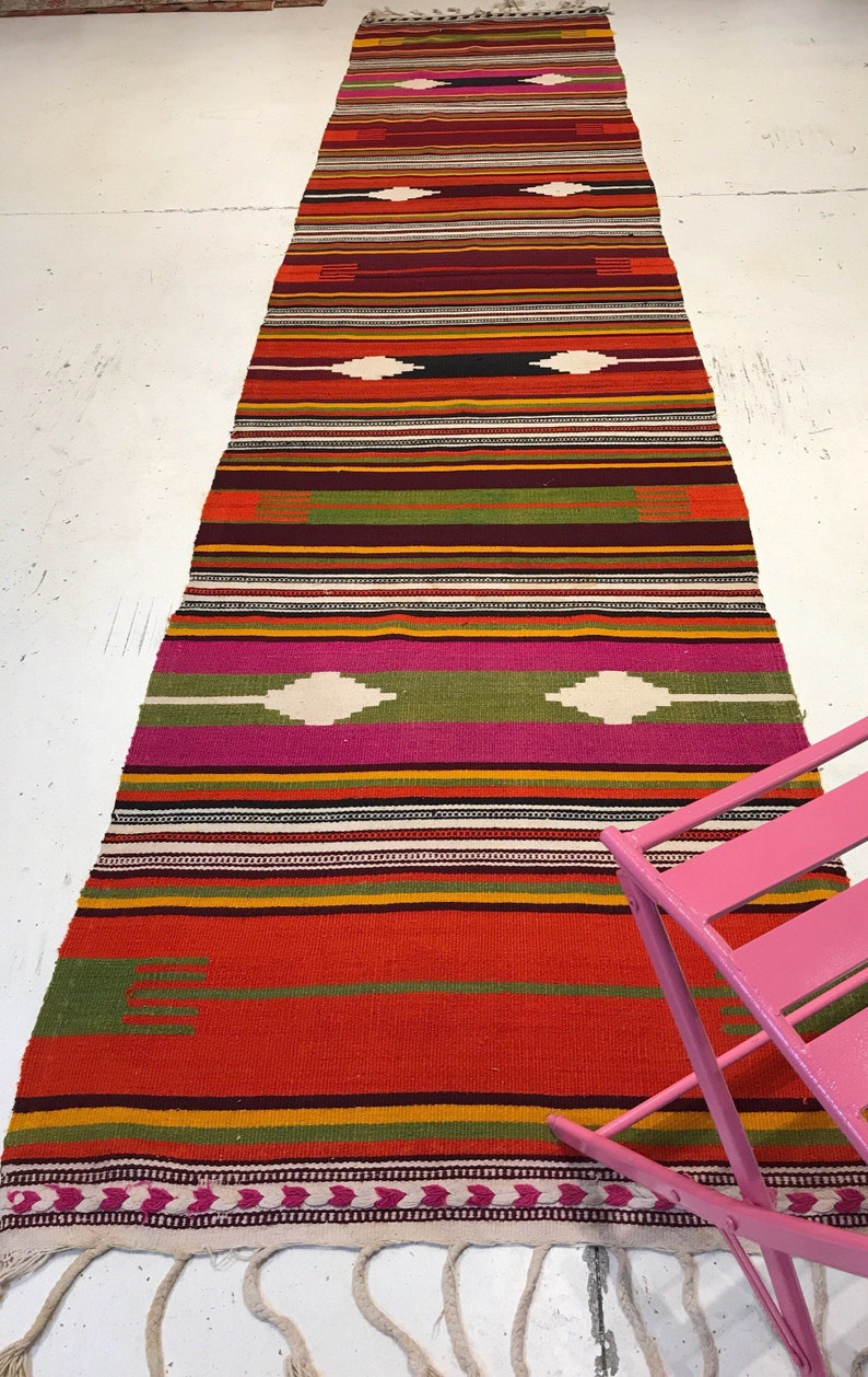 May include: A long, colorful rug with a striped pattern and geometric designs in shades of red, orange, green, pink, and white. The rug has fringe on the ends.