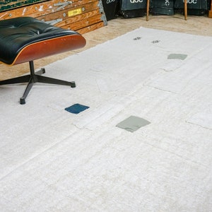 May include: A white, fluffy rug with a geometric design featuring squares of different colors, including blue and gray. The rug is laid out on a wooden floor with two black leather chairs with wooden bases.