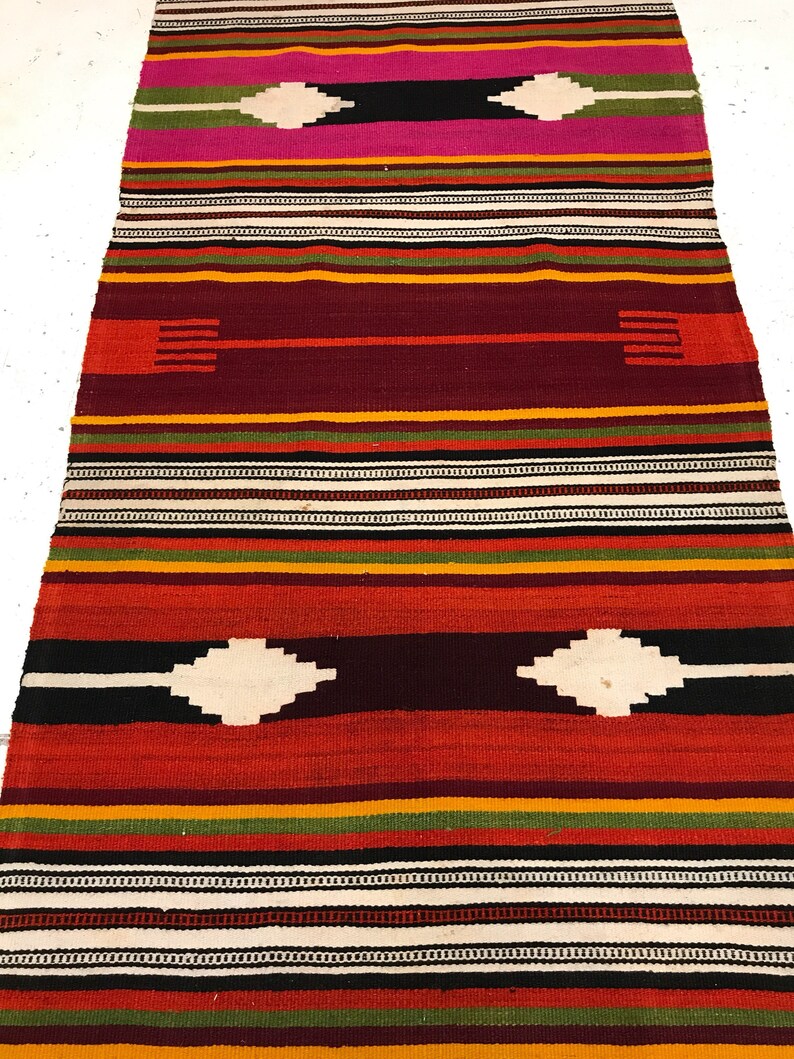 May include: A colorful striped rug with a geometric pattern. The rug is made of woven wool and has a variety of colors, including red, orange, yellow, green, black, and white.