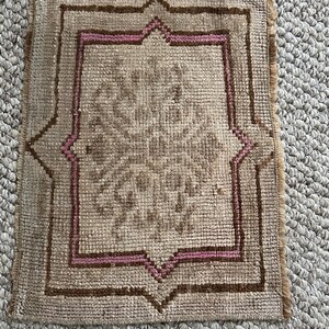 May include: A small, square rug with a brown and pink geometric pattern. The rug has a fringe border.