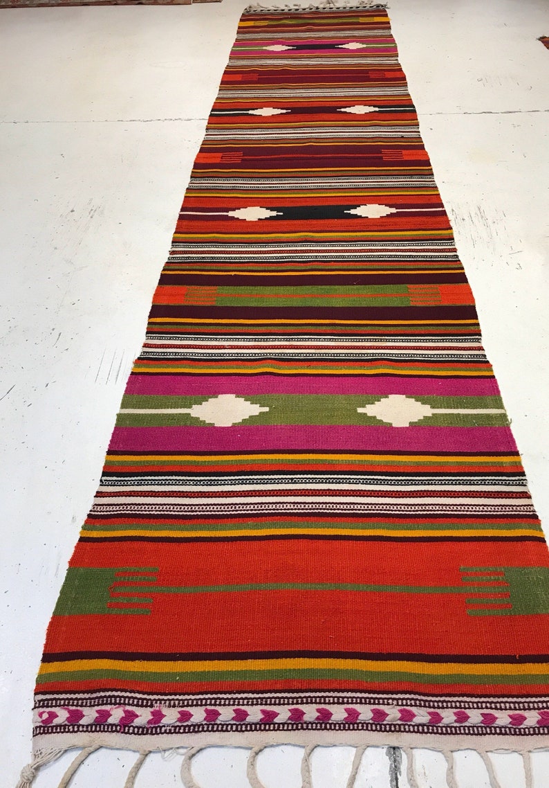 May include: A long, narrow, multi-colored rug with a geometric pattern. The rug has stripes of red, orange, yellow, green, pink, and white. The rug has a fringe on the bottom.