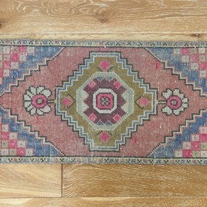 May include: A small, rectangular rug with a traditional Turkish design. The rug is primarily pink with a central medallion in shades of brown, green, and blue. The rug has a fringe border.