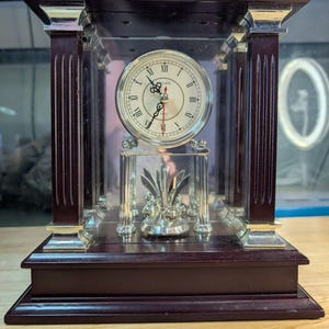 May include: A mantel clock featuring a dark wood frame with four silver-toned columns and a glass enclosure. The clock face is white with black Roman numerals and ornate black hands. A silver-toned pendulum with four decorative spheres sits below.