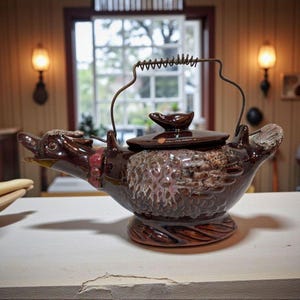 May include: A brown ceramic teapot shaped like a duck, with a detailed head, wings, and a handle. The teapot has a lid and sits on a decorative base. The background includes a window and warm lighting.