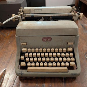 May include: Vintage Royal typewriter in a light gray color. The typewriter has a full keyboard with off-white keys and a carriage return lever. The Royal logo is visible on the front. The machine sits on a wooden surface.