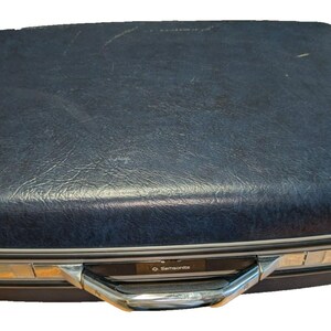 May include: A dark blue hard-shell suitcase with a textured finish. The rectangular case features a silver-toned metal trim along the center seam, a sturdy silver handle, and a small rectangular plate with the Samsonite brand name printed on it.