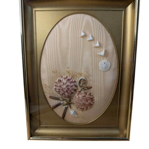 May include: Framed oval artwork featuring a beige fabric background with seashells and decorative elements. The frame is gold-colored. The artwork includes a variety of seashells, a starfish, and small white decorative pieces.