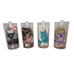 May include: Four plush animal toys, including a cat and three bunnies, are displayed in clear plastic containers. The cat wears a blue outfit, while the bunnies are dressed in pink, blue, and purple outfits. Each toy has a unique design.
