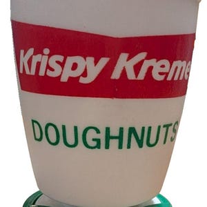 May include: A white plastic cup with a green rim and base. The cup features a red rectangular logo with the white text Krispy Kreme and the green text DOUGHNUTS printed below the logo. The cup has a tapered shape and a solid white body.