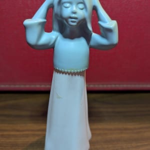Puede incluir: Una figurita de cerámica azul de una niña con el pelo largo, que lleva un vestido azul y una falda blanca. Se está sujetando las manos a la cabeza.