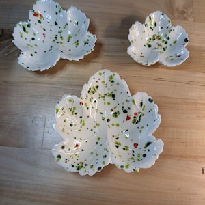 May include: Three white ceramic dishes shaped like maple leaves, each featuring a speckled pattern of green, red, and dark brown paint splatters across the surface. The dishes vary in size, with one large leaf-shaped bowl and two smaller ones.