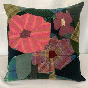 Puede incluir: Cojín decorativo cuadrado con diseño de patchwork. El cojín presenta un gran diseño floral rosa y rojo, con otros elementos florales en tonos morados, verdes y azules. El cojín está hecho de varias telas, incluyendo texturas a cuadros y lisas.