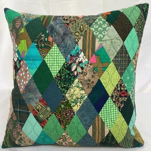 May include: Square cushion with a patchwork design. The cushion features a variety of green fabrics, including solid, patterned, and textured materials, arranged in a diamond pattern. The colour palette includes shades of teal, olive, and forest green.