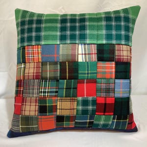 May include: Square decorative pillow with a patchwork design. The top and bottom sections are green plaid, while the center features a variety of plaid squares in red, blue, green, and tan. The pillow has a dark blue border.