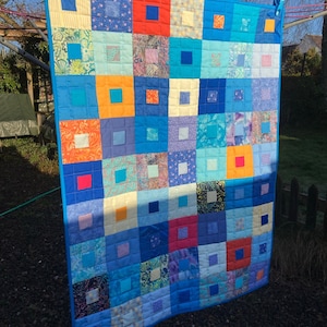 Può includere: Una trapunta patchwork blu e bianca con quadrati di diversi colori e motivi, tra cui arancione, rosso e giallo. La trapunta è appesa a uno stendino all'aperto.