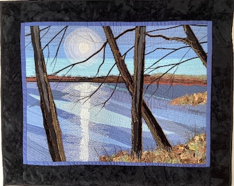 May include: A quilted wall hanging depicting a landscape scene with a lake, trees, and a bright moon in the sky. The lake is a light blue color and the trees are dark brown. The moon is a white circle with a light blue halo.