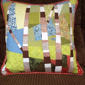 May include: A patchwork cushion with a geometric design featuring brown, green, white, blue, and red fabric squares. The cushion has a red border.