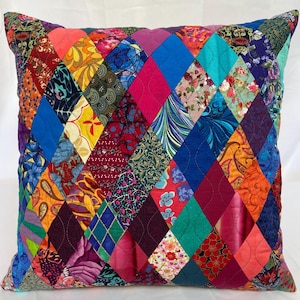 May include: Square decorative pillow with a patchwork design. The pillow features a diamond pattern with various fabric patterns and colors, including blue, red, orange, and floral prints. The pillow is quilted and has a soft, textured appearance.