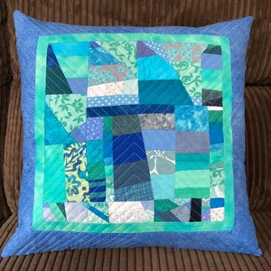 Op de afbeelding: Een blauw en groen patchwork kussen met een blauwe rand. Het kussen is gemaakt van verschillende stoffen met verschillende patronen.