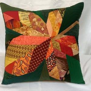 Könnte beinhalten: Quadratisches Zierkissen mit Patchwork-Design. Das Kissen hat ein zentrales Blumenmuster in Orange-, Rot-, Gelb- und Brauntönen auf einem dunkelgrünen Hintergrund. Der Patchwork umfasst verschiedene Stoffmuster und -texturen.