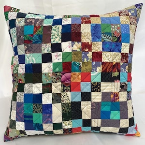 May include: Square patchwork cushion featuring a variety of fabric patterns and colours, including blue, green, red, and floral designs. The cushion is quilted with a grid pattern, creating a textured surface. The cushion is a square shape.