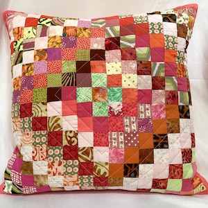 May include: A patchwork cushion with a pink background and squares of various colours and patterns, including green, brown, orange, and pink.