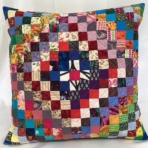 Puede incluir: Cojín cuadrado con diseño de patchwork. El cojín presenta un patrón de cuadrícula de pequeños cuadrados de tela coloridos en varios patrones y tonos, incluyendo estampados rojos, azules, verdes y florales. El cojín tiene una apariencia acolchada.