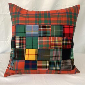 May include: Square cushion with a patchwork design. The cushion has a red and green tartan pattern on the top and bottom, with a patchwork of various tartan patterns in the centre. The cushion is square and appears to be made of fabric.